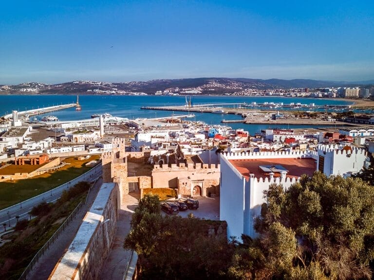 Veduta aerea della città costiera di Tangeri con uno storico castello in primo piano e un porto turistico con barche ed edifici moderni sullo sfondo, sullo sfondo di colline e un cielo azzurro e limpido. Parte della Città Imperiali, è una tappa essenziale in qualsiasi tour del Marocco Classico.