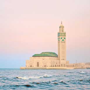 Una moschea con una cupola verde e un alto minareto si erge adiacente al mare, sotto un cielo rosa tenue, catturando l’incantevole fascino spesso evidenziato nell’offerta viaggio in Marocco di SBS Viaggi.