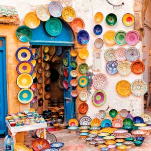 L'ingresso di un negozio adornato con piatti di ceramica colorati e dai motivi intricati allude all'offerta viaggio in Marocco di SBS Viaggi. Altri piatti sono esposti su un tavolo e coprono le pareti circostanti.