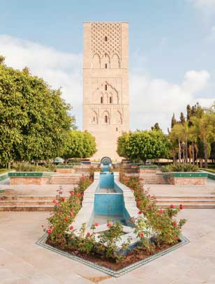 L'immagine mostra la Torre Hassan a Rabat, in Marocco, con un gioco d'acqua e un giardino in primo piano, incorniciata da alberi su entrambi i lati sotto un cielo azzurro. Scopri questo scenario mozzafiato attraverso la nostra offerta viaggio in Marocco con SBS Viaggi.