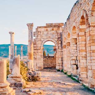 Antiche rovine in pietra con diverse colonne, un arco e mura parziali, sullo sfondo di colline e un cielo limpido: esplora queste meraviglie senza tempo nella tua prossima offerta viaggio in Marocco con SBS Viaggi.