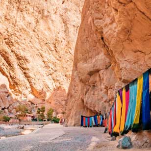 Alte pareti del canyon con tessuti colorati appesi lungo un lato. Sullo sfondo sono visibili il terreno roccioso e un po' di verde, una scena perfetta per un'avventura di SBS Viaggi offerta viaggio in Marocco.