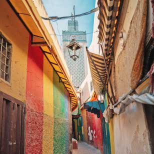 Uno stretto vicolo con edifici colorati e tende a strisce conduce a un'alta torre decorata della moschea sullo sfondo, che ricorda le scene di un SBS Viaggi offerta viaggio in Marocco.