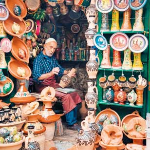 Un uomo siede in un negozio circondato da ceramiche colorate e lavora su un vaso di terracotta che tiene in grembo. Sugli scaffali e appesi intorno a lui sono esposti vari pezzi di ceramica e terracotta decorati, che ricordano i tesori che si potrebbero scoprire con un'offerta viaggio in Marocco di SBS Viaggi.