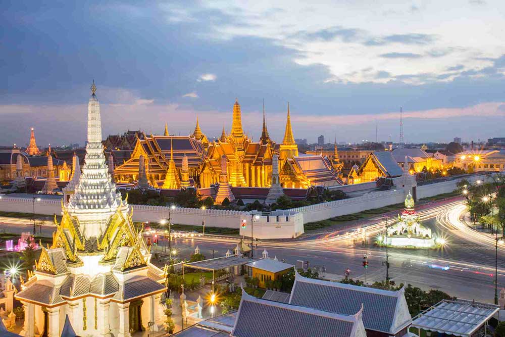 Veduta del complesso illuminato del Grand Palace al tramonto a Bangkok, con edifici decorati, templi e una strada piena di auto in primo piano: una scena affascinante che ti aspetta con l'offerta viaggio in Thailandia di SBS Viaggi.