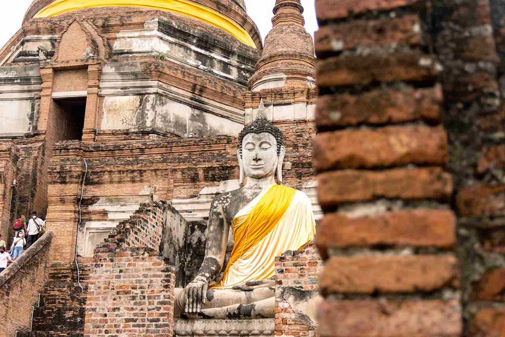 Una grande statua di Buddha seduto, avvolta in un panno giallo, si erge maestosa di fronte ad un antico tempio in mattoni e pietra, uno spettacolo da non perdere con l'offerta viaggio in Thailandia di SBS Viaggi.