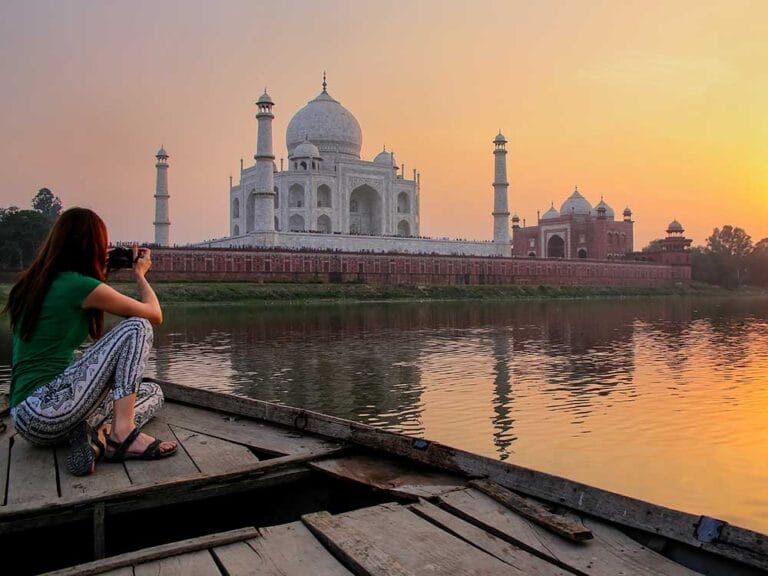 Persona su una barca di legno che fotografa il Taj Mahal attraverso un fiume al tramonto, catturando le emozioni di questo viaggio in India.