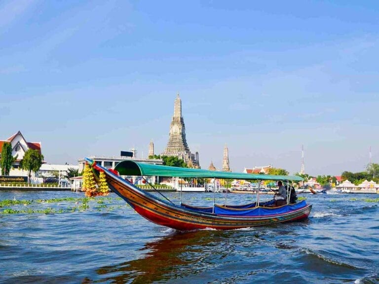 Una colorata barca a coda lunga viaggia sul fiume Chao Phraya con il tempio Wat Arun visibile sullo sfondo sotto un cielo azzurro, mostrando l'autentica Thailandia.