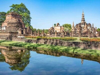 Le rovine dell'antico tempio con strutture in mattoni e pilastri in pietra, riflesse in un calmo specchio d'acqua e circondate da una vegetazione lussureggiante sotto un cielo azzurro e limpido, evocano lo splendore classico della Thailandia.