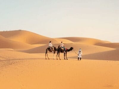 Due persone cavalcano cammelli attraverso un deserto mentre una persona vestita di bianco le guida, mettendo in mostra la bellezza di Tutto Marocco. Il paesaggio presenta ampie dune di sabbia sotto un cielo limpido, catturando l'essenza di questo incantevole viaggio marocchino.