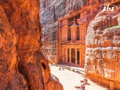 Una veduta di Al-Khazneh, l'edificio del Tesoro, a Petra, in Giordania, scolpito nelle scogliere di roccia rossa. Davanti alla struttura sono visibili diversi cammelli e persone.
