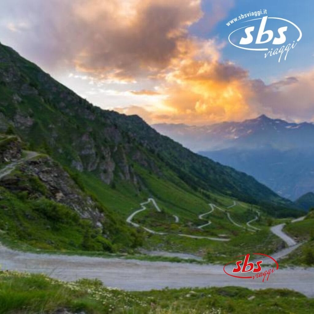 Strada di montagna tortuosa con vegetazione lussureggiante e nuvole spettacolari al tramonto, che conduce allo straordinario TH Sestriere. Logo 