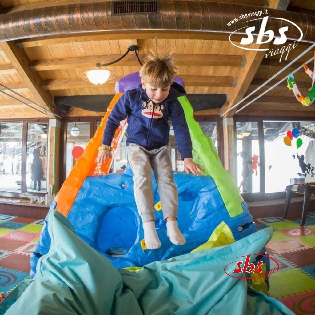 Un bambino con una felpa blu salta da uno scivolo gonfiabile all'interno di un'area giochi al coperto a La Thuile, decorata con tappeti colorati e palloncini.