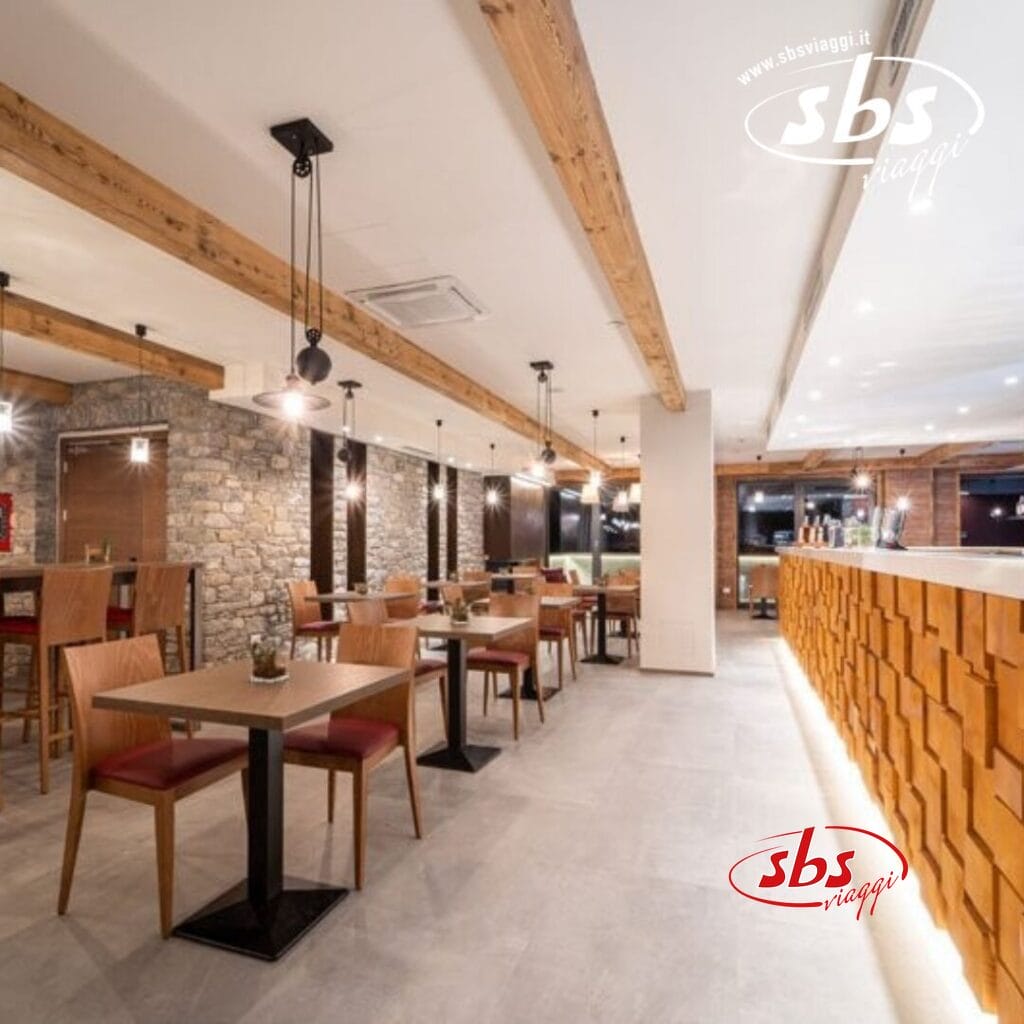 Un moderno ristorante interno con mobili in legno, travi a vista, pareti in pietra e un'elegante area bar. L'ambiente è ben illuminato con luci pendenti e presenta un logo nell'angolo in alto a destra che mostra con orgoglio TH Courmayeur.