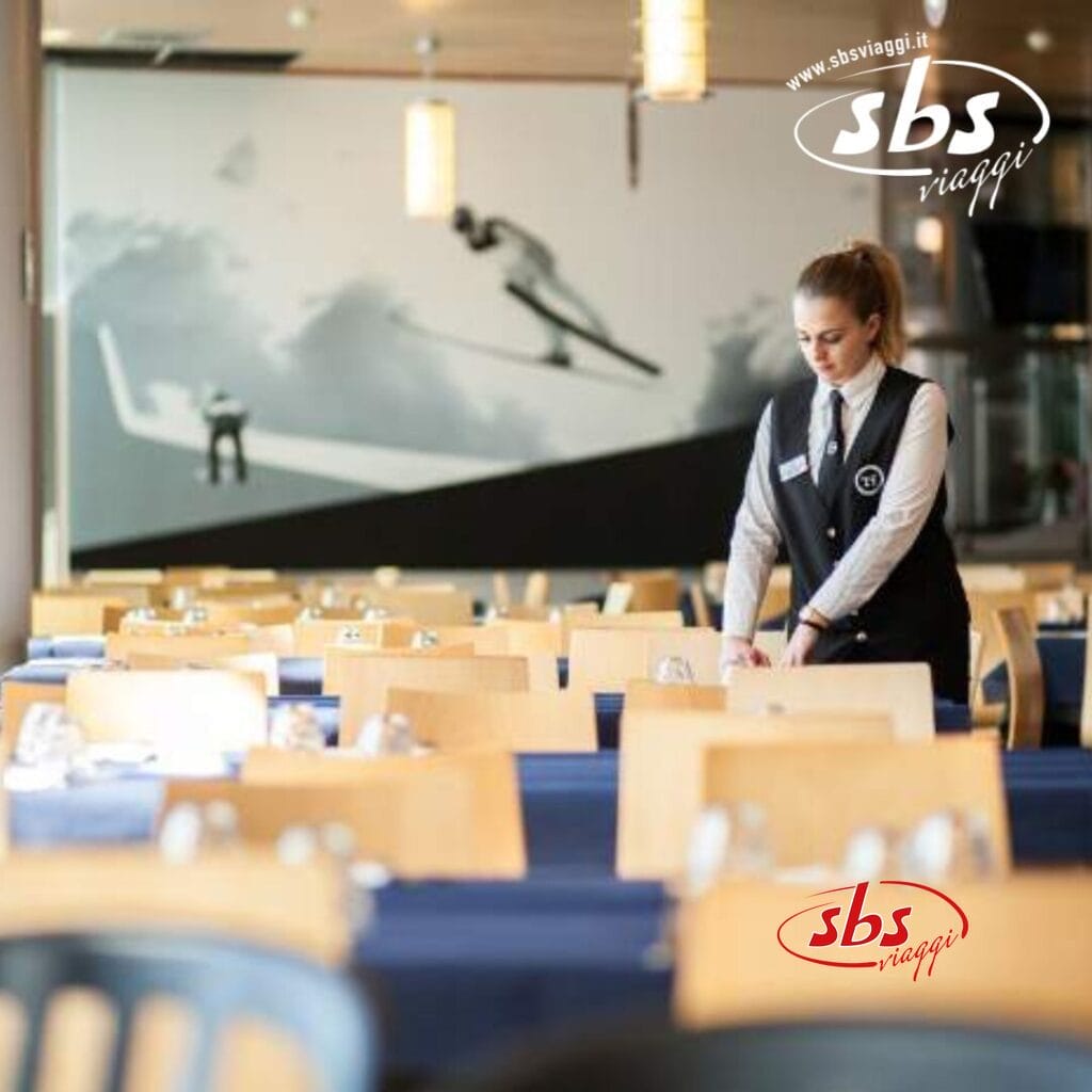 Una cameriera apparecchia i tavoli in un ristorante con tovaglie blu al Villaggio Olimpico. Un murale di sciatori decora la parete di fondo, che ricorda il TH Sestriere. Il logo 