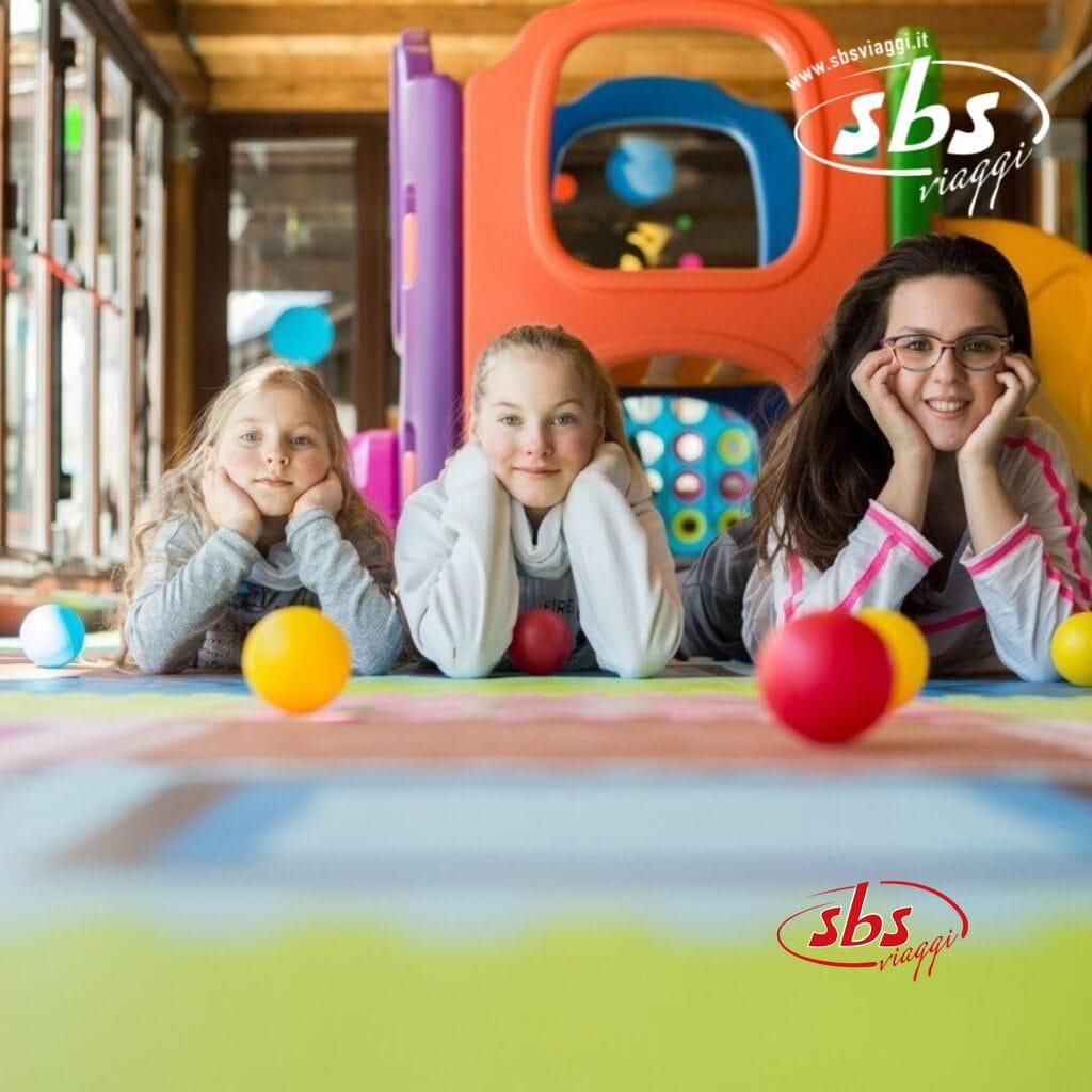 Tre bambini giacciono a pancia in giù, sorridenti, con il mento tra le mani su un tappetino da gioco colorato. Una struttura di gioco vivace e palline di plastica creano uno sfondo gioioso; il logo 