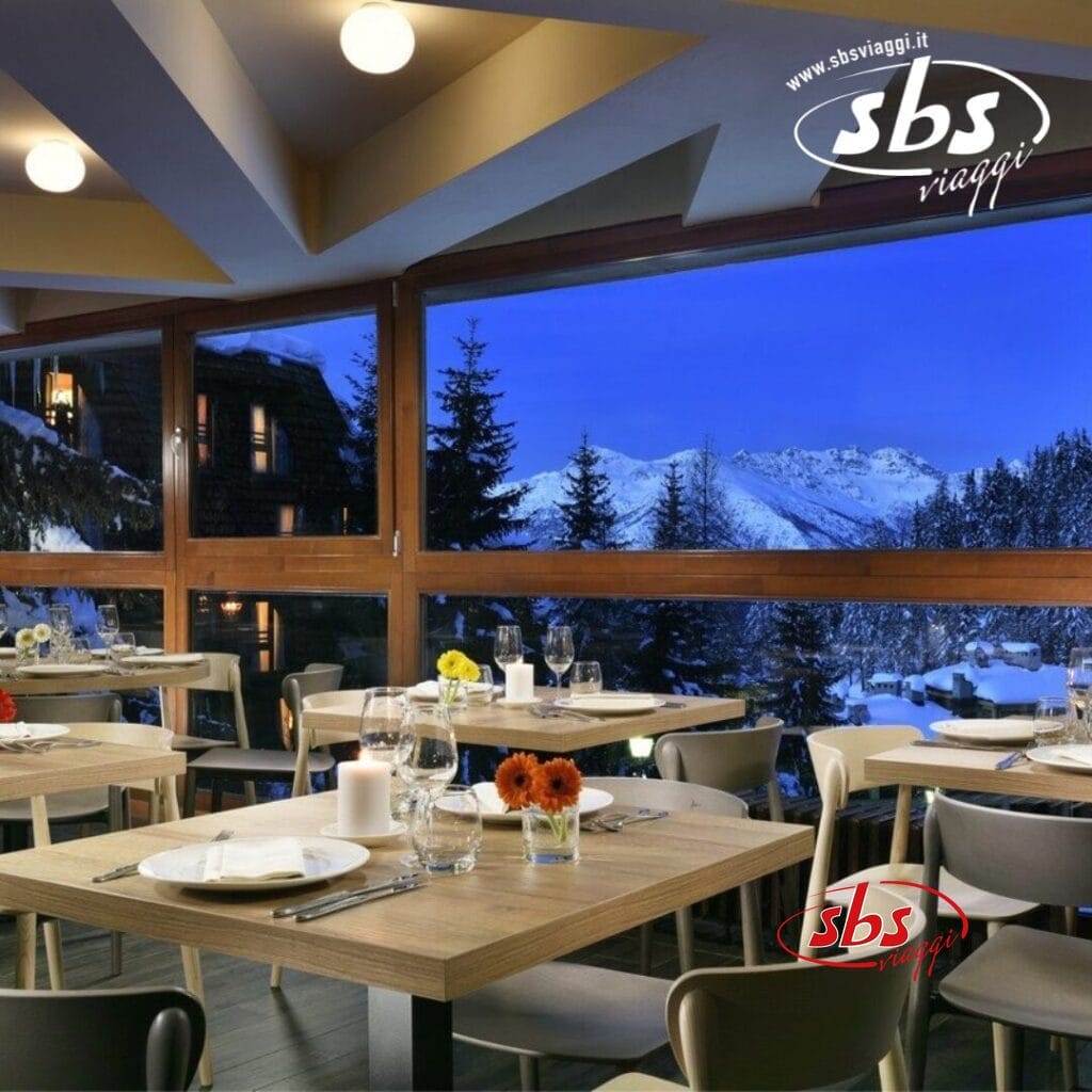 Ristorante moderno con tavoli in legno, ordinatamente apparecchiati con piatti, bicchieri e centrotavola floreali. Ampie finestre offrono una vista panoramica sulle montagne innevate e sui pini al tramonto, creando un'atmosfera incantevole, perfetta per cenare al Pila.