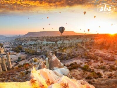 Un paesaggio all'alba del 2025 mostra numerose mongolfiere che fluttuano sopra un terreno roccioso e desertico con formazioni rocciose uniche e altipiani lontani, catturando l'essenza del Tour Della Turchia. Vicino all'orizzonte, uno storico Kervansaray aggiunge misticismo a questa scena mozzafiato.