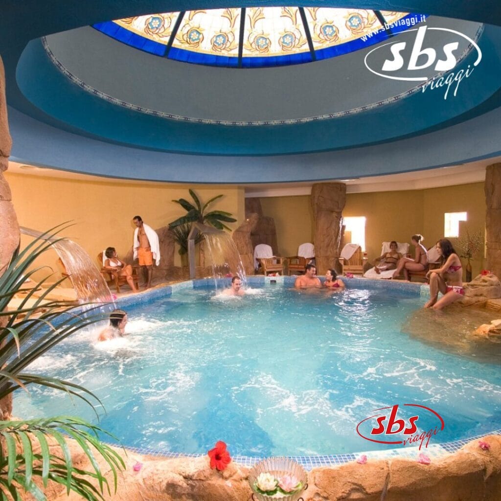 Persone che si rilassano e si divertono in una piscina coperta del Caribbean World Resort, Soma Bay. La scena comprende formazioni rocciose decorative, un soffitto a cupola in vetro colorato, giochi d'acqua e piante tropicali.