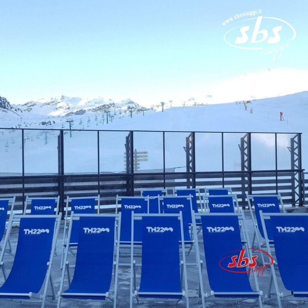 File di sedie a sdraio blu su una terrazza innevata al Th La Thuile si affacciano su una pista da sci con seggiovie sullo sfondo. Sulle sedie è stampato 