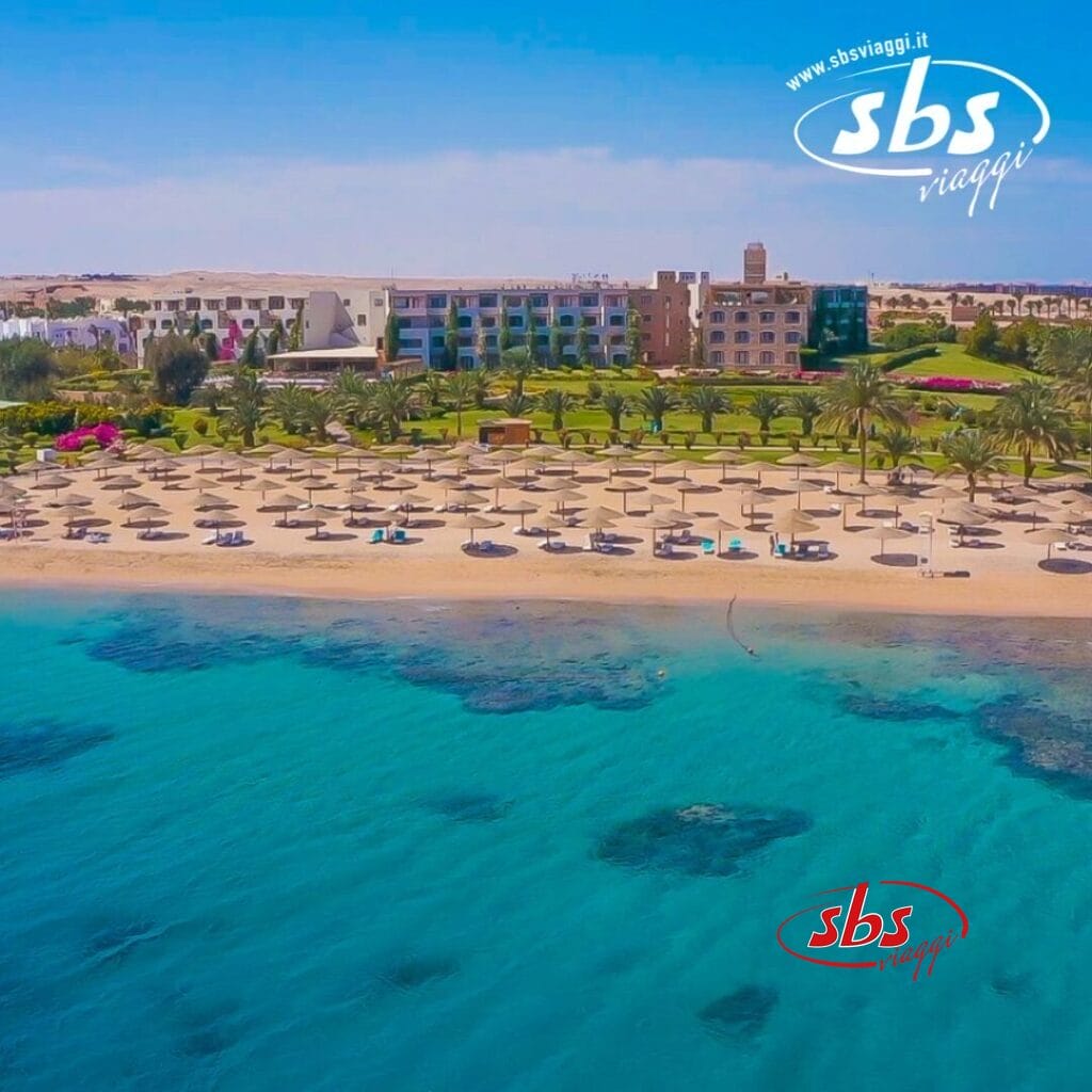 Vista aerea del Fort Arabesque Resort con ombrelloni e lettini sulla sabbia, mare turchese e un edificio dell'hotel sullo sfondo. Sono disponibili strutture spa per il relax. Il logo 