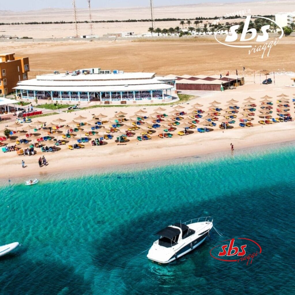 Vista aerea di Soma Bay con un resort sulla spiaggia con file di ombrelloni colorati e lettini, un edificio bianco e una barca ancorata in acque azzurre e limpide. Sullo sfondo sono visibili terreni marroni e strutture secondarie.