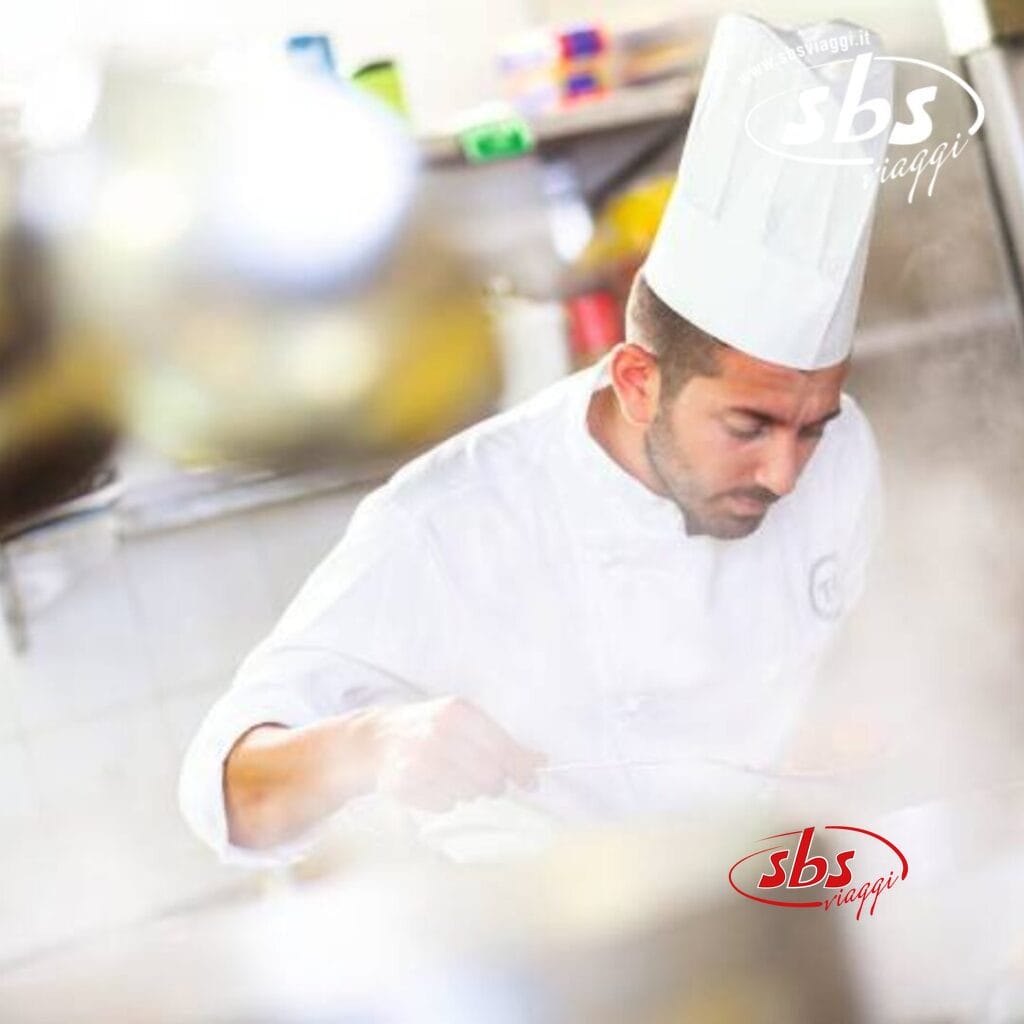 Uno chef in uniforme bianca e con un alto cappello da cuoco cucina in una cucina affollata e piena di vapore al Villaggio Olimpico di Sestriere.