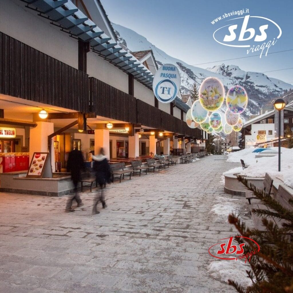 Scena di strada invernale a La Thuile con terreno innevato ed edifici in legno decorati con ornamenti a tema pasquale. Due persone camminano e un cartello di un negozio recita "Buona Pasqua". Maestose montagne si ergono alte sullo sfondo.