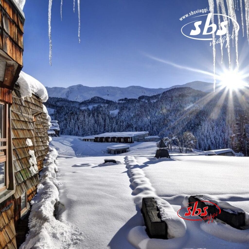 Paesaggio innevato con baite, sentieri innevati, ghiaccioli e montagne lontane sotto un sole splendente. Il logo Pila nell'angolo in alto a destra recita 