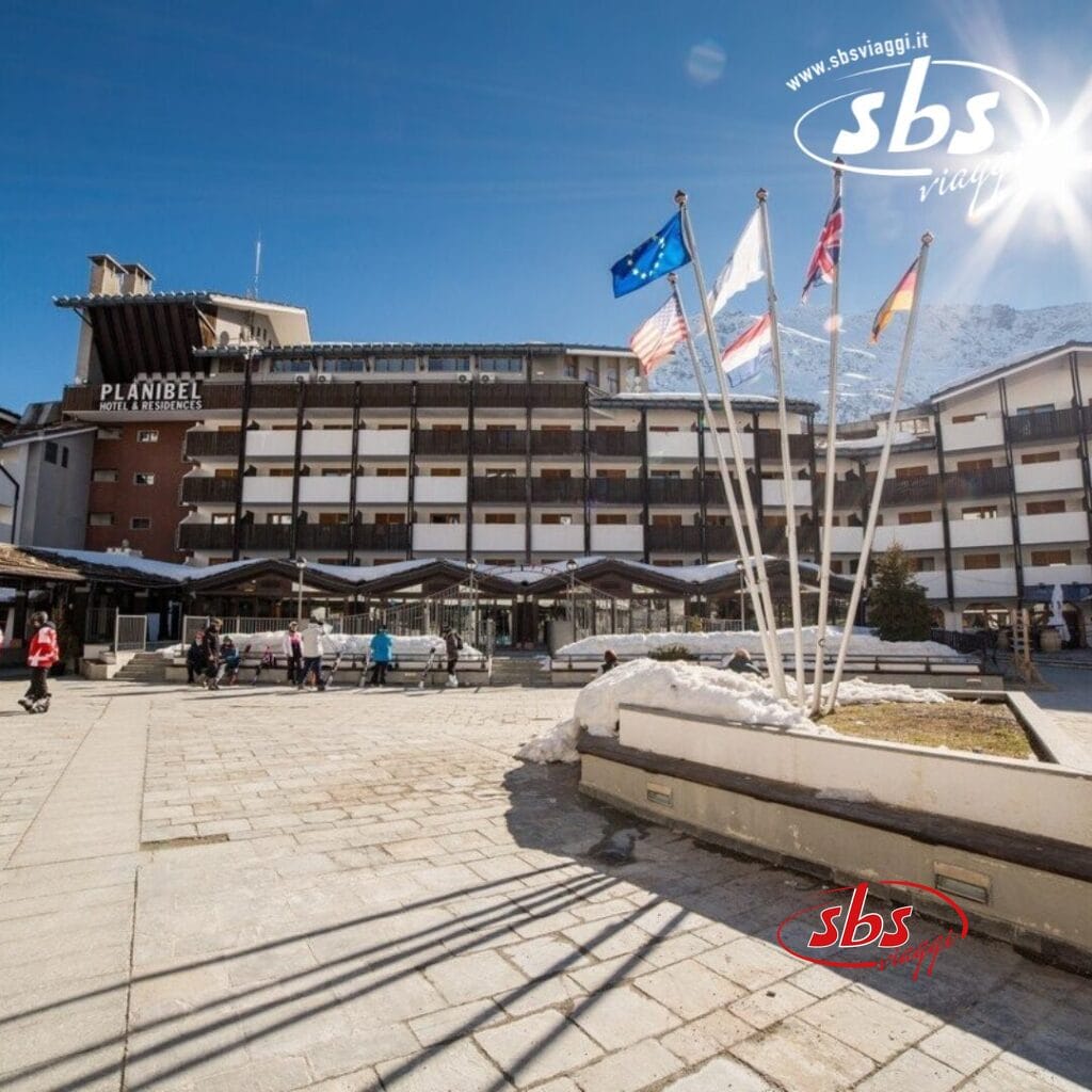 Un hotel con più bandiere all'ingresso e una montagna sullo sfondo, incastonato nell'incantevole cittadina di La Thuile. Le persone camminano nel cortile di questo edificio Planibel sotto un cielo terso e soleggiato.