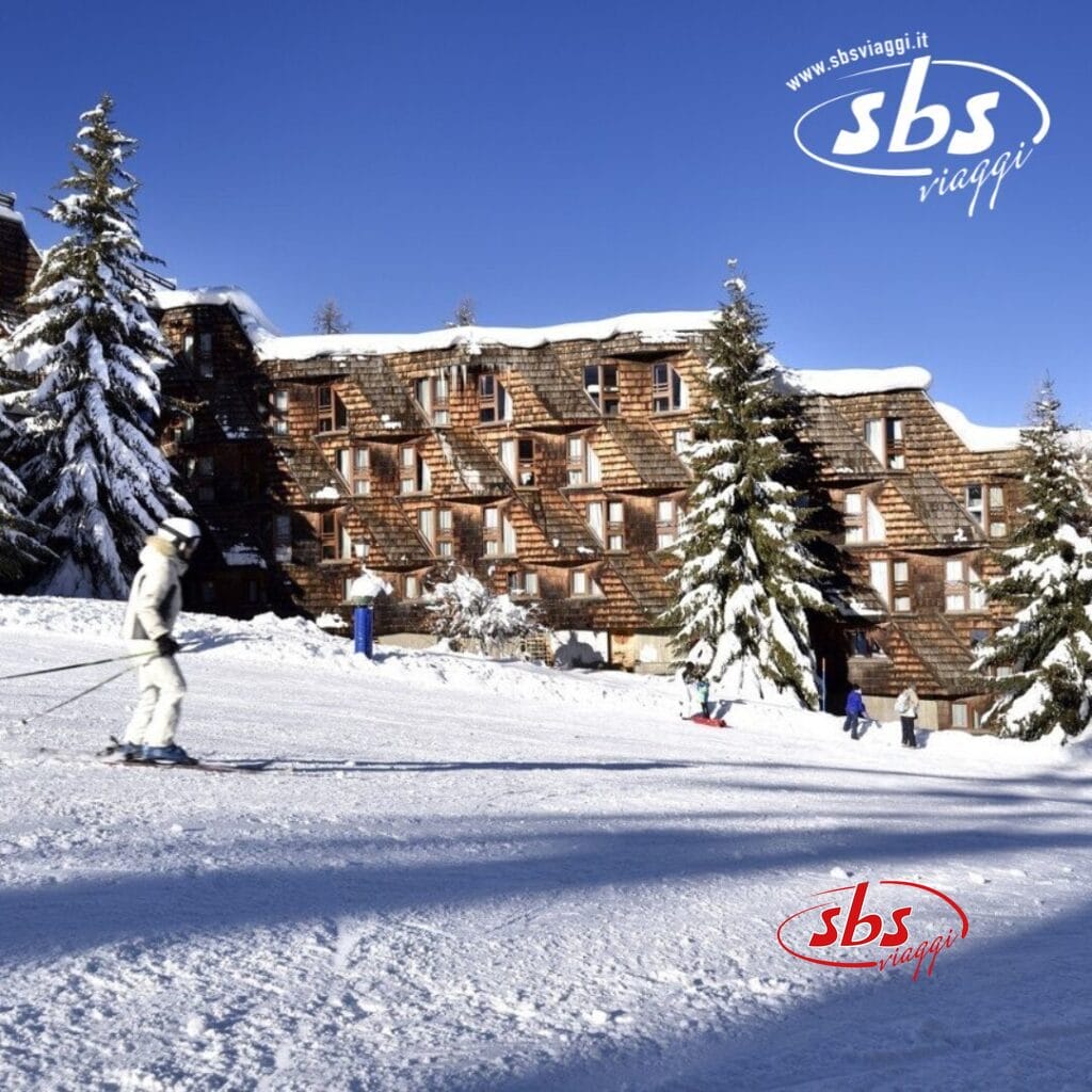 Sciatori su un pendio innevato di fronte a un edificio in stile chalet con alberi innevati, sotto il cielo azzurro e terso di Th Pila. Un logo 