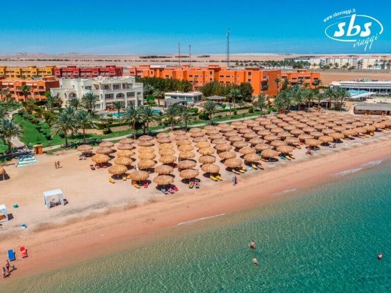 Vista aerea del Caribbean World Resort a Soma Bay, che mostra file di ombrelloni di paglia e sedie a sdraio sulla riva sabbiosa. Edifici, palme e vegetazione lussureggiante sono visibili sullo sfondo, incarnando il lusso tropicale.