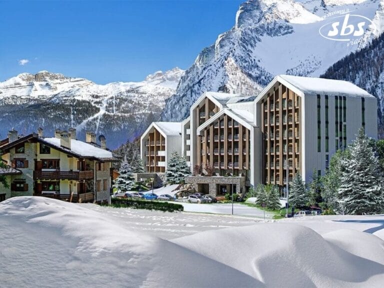 Immagine del complesso alberghiero TH Courmayeur con tetti innevati e montagne sullo sfondo. Un parcheggio con auto e qualche albero sono visibili di fronte agli edifici.