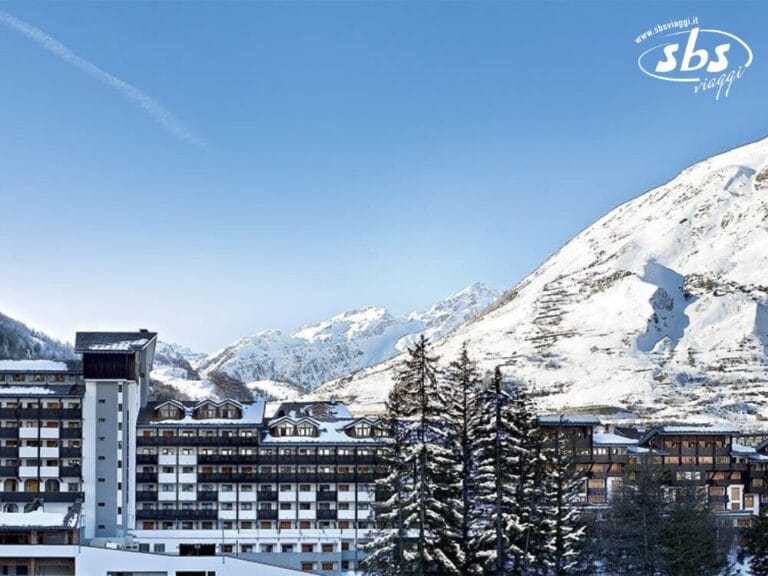 Una località di montagna innevata con numerosi edifici, alberi in primo piano e un cielo azzurro e limpido a La Thuile. Nell'angolo in alto a destra è scritto 