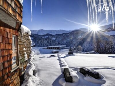 Una baita in legno circondata dalla neve con una vista nitida sulle Alpi italiane e un paesaggio montano innevato. La luce del sole splende intensamente, proiettando ombre, mentre grandi ghiaccioli pendono dal bordo dell'edificio a Th Pila.