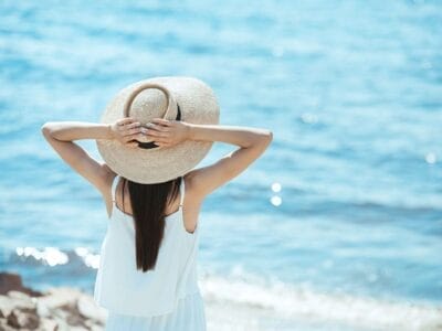 Persona che indossa un abito bianco e un cappello di paglia, in piedi con le spalle alla telecamera, le mani sul cappello, che ammira il mare tranquillo durante la sua vacanza.