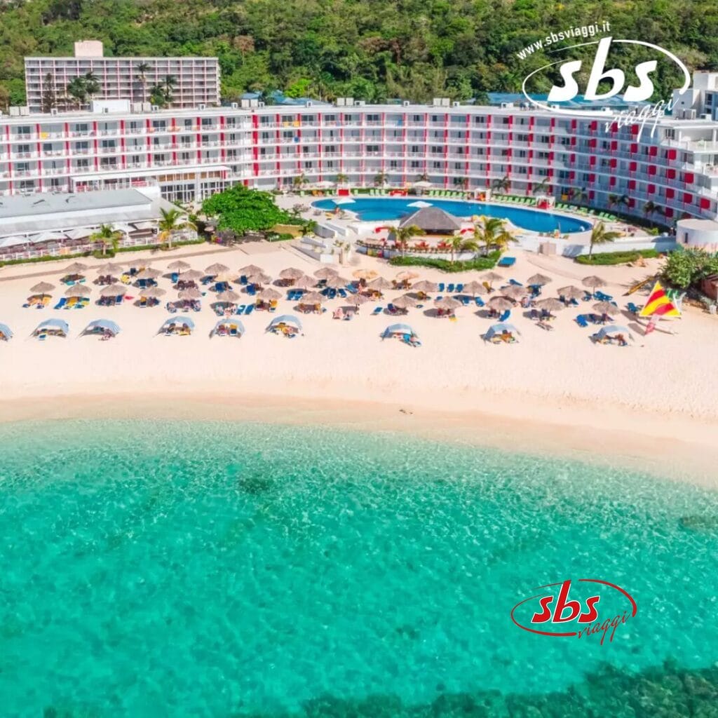 Vista aerea di un resort sulla spiaggia, caratterizzato da un hotel in stile bozza automatica, una piscina scintillante e file di ombrelloni e sedie a sdraio sulla spiaggia sabbiosa, il tutto sullo sfondo di un'acqua turchese e limpida.