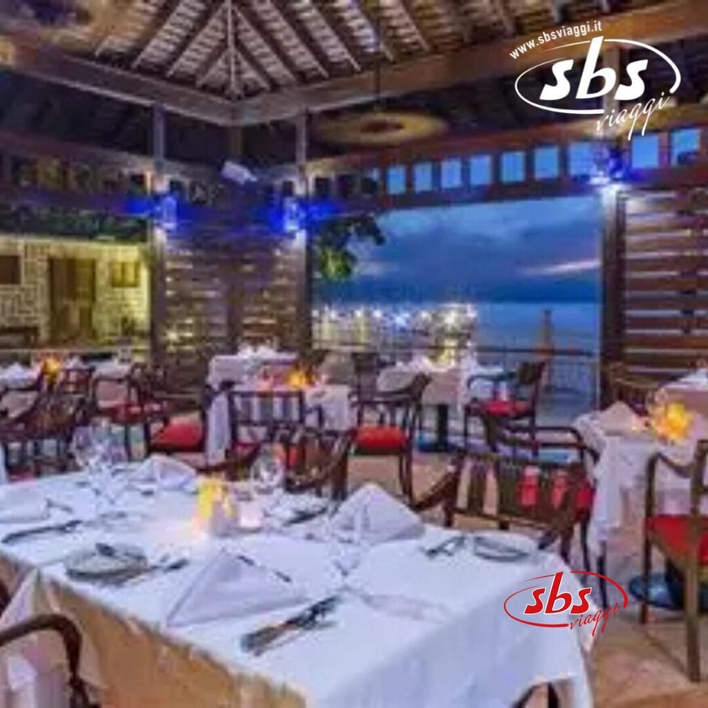 Al Royal Decameron Montego Beach, goditi un ristorante all'aperto con tavoli ordinatamente apparecchiati che si affacciano sul lungomare al tramonto. L'arredamento in legno e l'illuminazione calda creano un'atmosfera accogliente, perfetta per gli ospiti di età superiore ai 16 anni. Il logo 