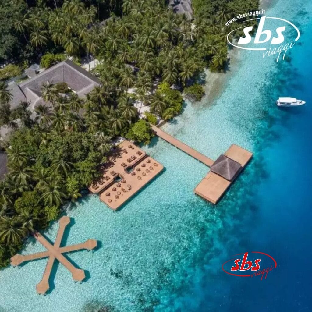 Vista aerea di un resort tropicale, che ricorda Fihalhohi, con passerelle e strutture sull'acqua. Circondato da acque azzurre e lussureggianti, e da una barca nelle vicinanze, questo ambiente pittoresco racchiude il fascino del paradiso del Club Coralia.