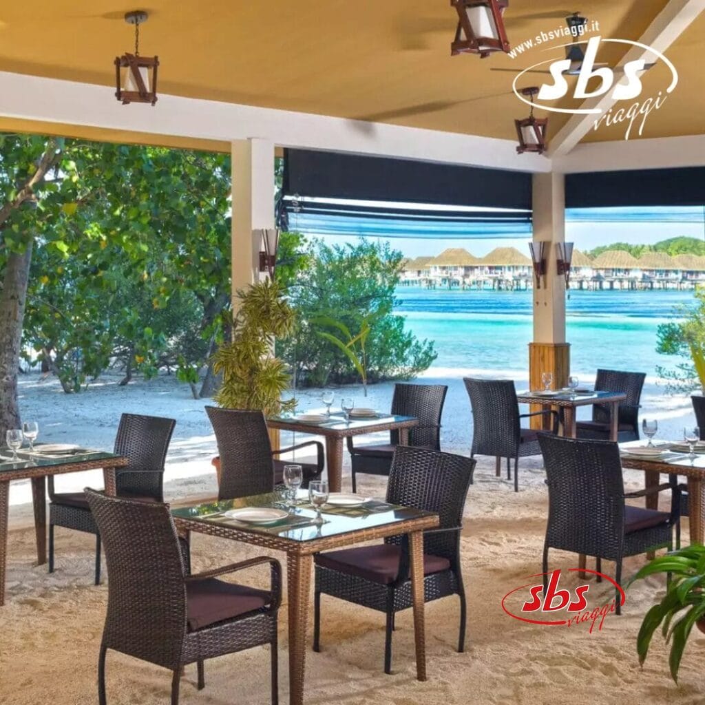 La zona pranzo all'aperto del Pearl Sands è dotata di sedie in vimini e tavoli quadrati su un pavimento sabbioso, con vista su una splendida spiaggia tropicale. L'ambiente del boutique hotel comprende acque limpide e affascinanti strutture con tetti di paglia sullo sfondo.