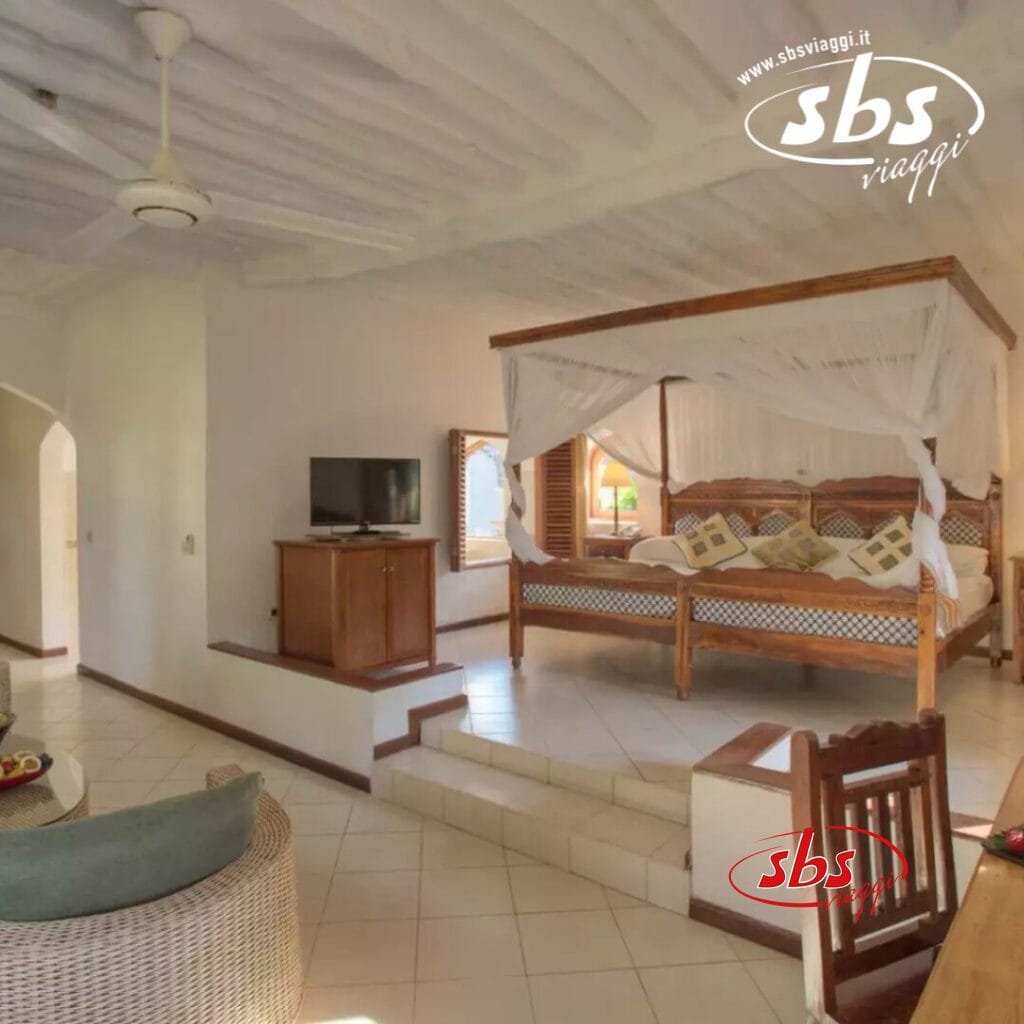 Una camera da letto accogliente al Bluebay Beach Resort dispone di un letto a baldacchino, una TV su un supporto di legno e una sedia di vimini. Con pareti chiare e pavimento piastrellato, la stanza trasuda tranquillità. Un arco aperto offre un accesso senza soluzione di continuità a un altro spazio, riecheggiando l'atmosfera rilassante della spa del resort.