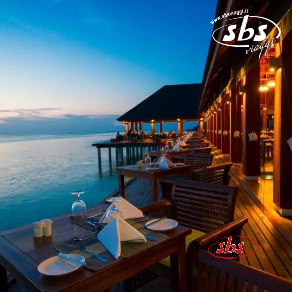 I tavoli da pranzo allestiti su una terrazza in legno con vista sull'oceano al tramonto evocano l'essenza di un'idilliaca serata estiva, con un affascinante edificio in legno che si estende sull'acqua, catturando perfettamente l'atmosfera rilassata dell'isola.