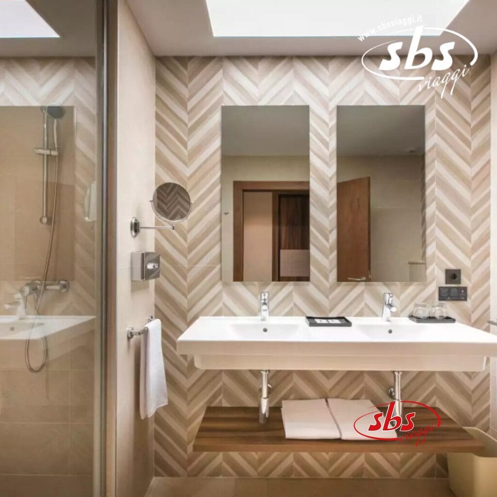 Questo moderno bagno del Riu Jambo Hotel è dotato di un elegante doppio lavabo, un grande specchio e pareti con motivi geometrici alla moda. Sulla sinistra, troverete una spaziosa cabina doccia che aggiunge fascino contemporaneo.