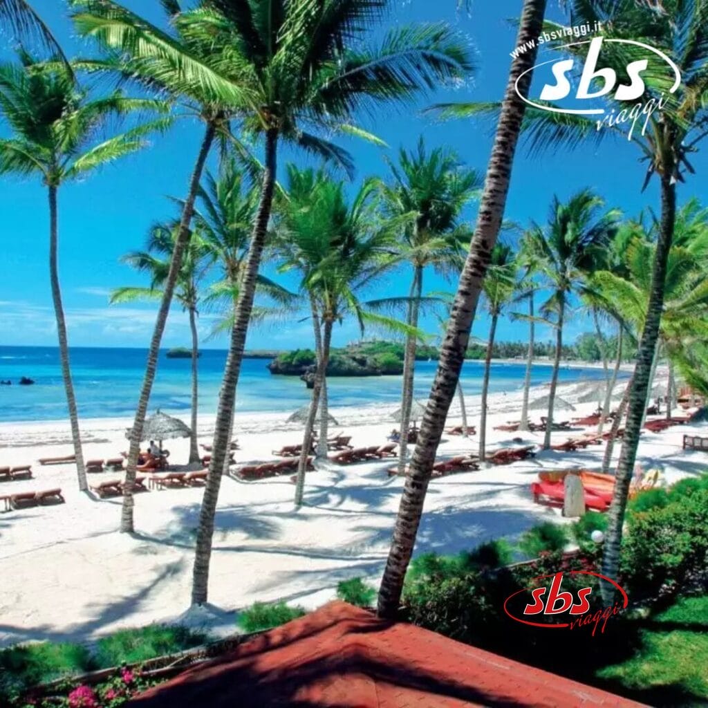 Scena di spiaggia con palme, sedie a sdraio e il mare azzurro e limpido di Crystal Bay sotto un cielo luminoso. Le strutture con i tetti rossi del resort Veraclub sono parzialmente visibili.