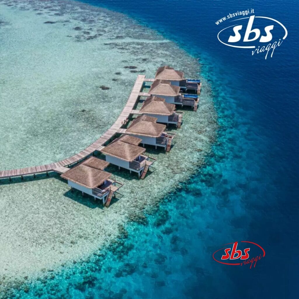 Vista aerea dei bungalow sull'acqua del Cocogiri Island Resort, affacciati su un limpido oceano azzurro, collegati tra loro da una passerella in legno, con il logo SBS Viaggi in un angolo.