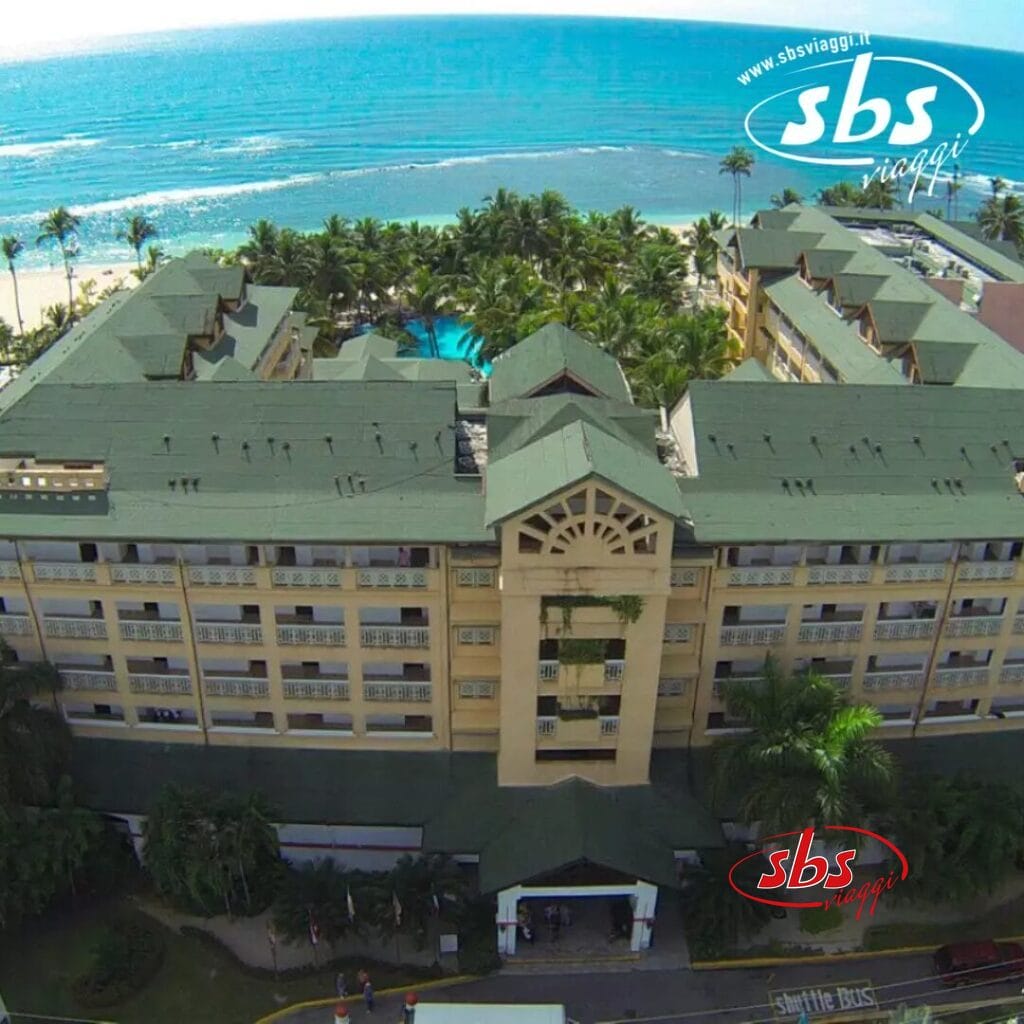 Una vista aerea del Coral Costa Caribe rivela un hotel sulla spiaggia con tetti verdi e lussureggianti, incastonato tra palme ondeggianti. Il vivace Beach Resort offre viste mozzafiato con l'oceano che si estende serenamente sullo sfondo.