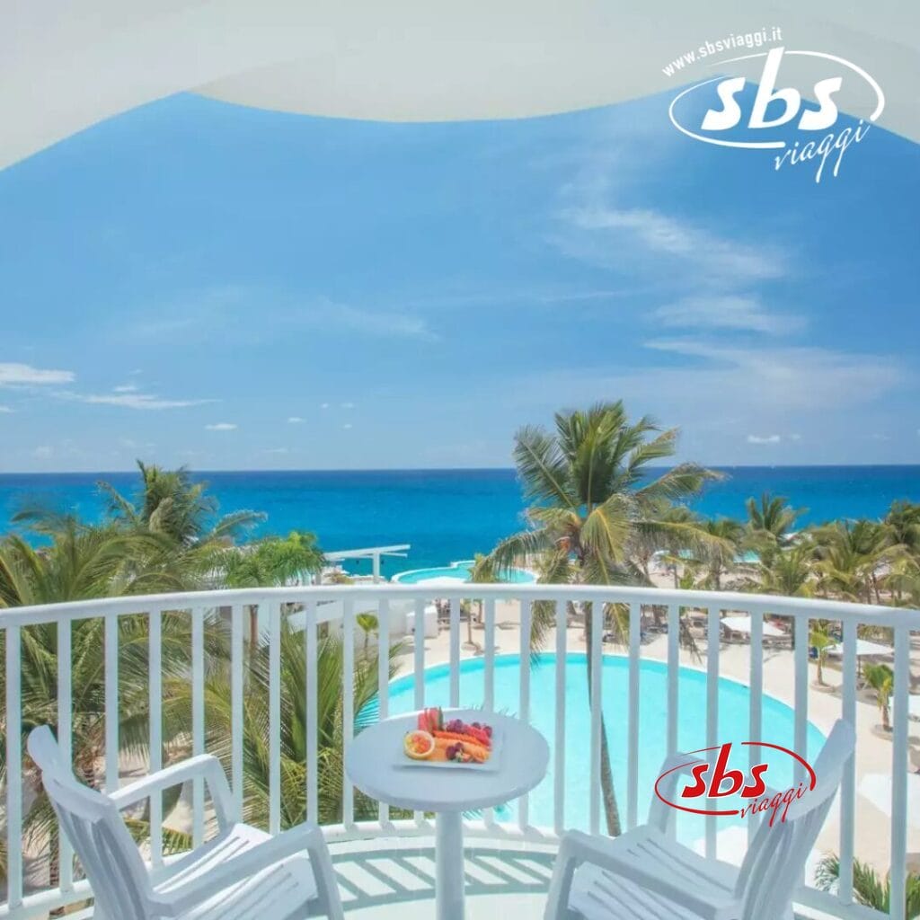 Dal balcone, godetevi la vista mozzafiato: una piscina tranquilla, palme ondeggianti e il vasto oceano sotto un cielo azzurro e limpido, il tutto incorniciato da due invitanti sedie e un grazioso tavolo in primo piano.