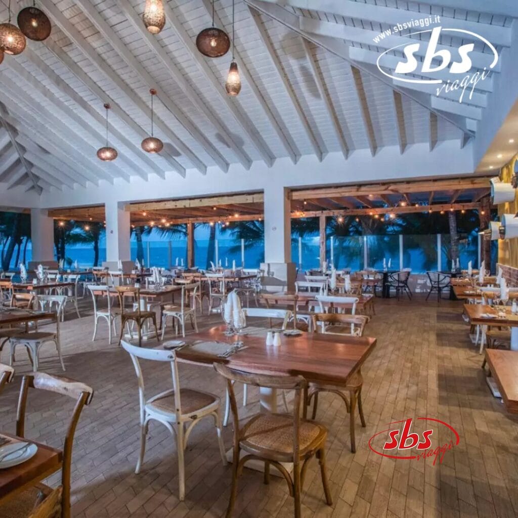 Un ristorante sulla spiaggia con tavoli e sedie in legno sotto un soffitto a volta bianco, con luci pendenti e vista sull'oceano attraverso pareti aperte. Noto per la sua atmosfera da bozza automatica, lo spazio invita al relax con dolci brezze marine che esaltano l'esperienza culinaria.