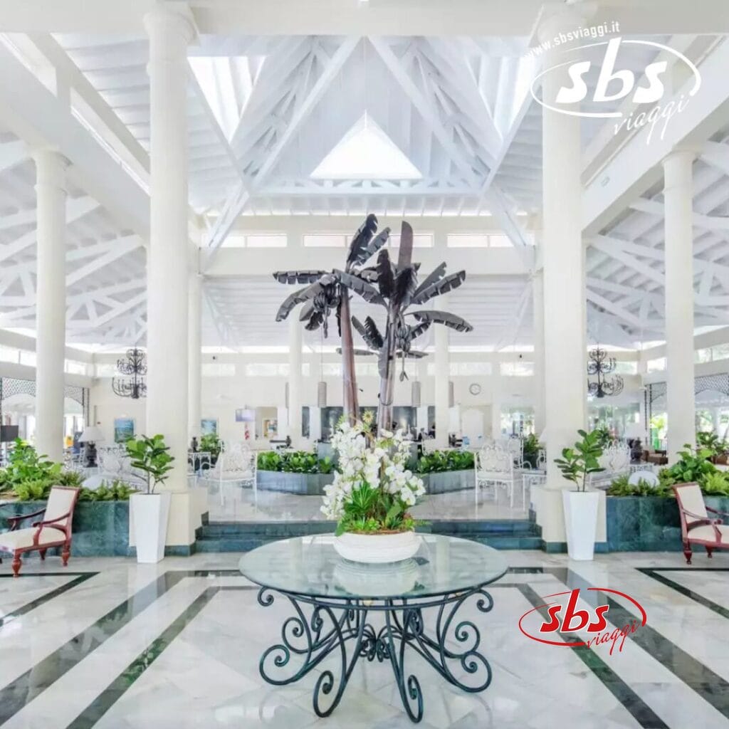 La spaziosa hall del Grand Bahia Principe presenta soffitti alti, pilastri bianchi e un tavolo centrale in vetro decorato con una pianta vivace. Le decorazioni con palme completano le eleganti aree salotto, creando un'atmosfera invitante che ricorda il fascino di El Portillo.