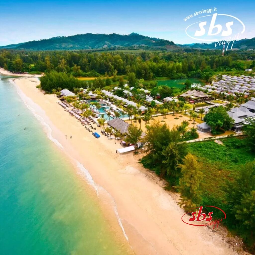 Vista aerea di un resort sulla spiaggia con vegetazione lussureggiante, edifici sparsi e sedie a sdraio vicino alla riva. Colline e alberi fitti sono visibili sullo sfondo, creando una fuga pittoresca per chi cerca tranquillità in mezzo alla bellezza della natura.